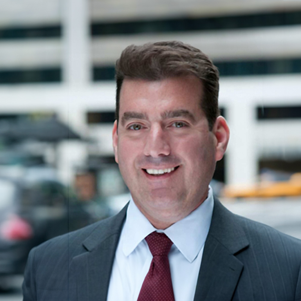 The image shows a man wearing a suit with a tie, standing indoors against a cityscape background.