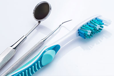 The image depicts a collection of dental hygiene tools, including a toothbrush with blue bristles, a dental mirror, and dental instruments such as forceps and possibly tweezers or other small tools, all laid out on a white surface.