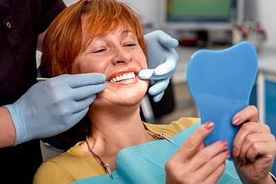 The image shows a woman sitting in a dental chair, smiling broadly with her eyes closed, while holding a blue object that appears to be a dental impression tray. A dental professional is standing behind her, adjusting the tray and wearing gloves.