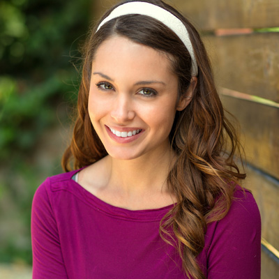 The image shows a young woman with long brown hair, wearing a purple top and a white headband, posing for the camera against a wooden fence background.