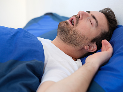 A man in a white shirt is lying down on a bed with a blue comforter, his eyes are closed and he appears to be sleeping or resting.